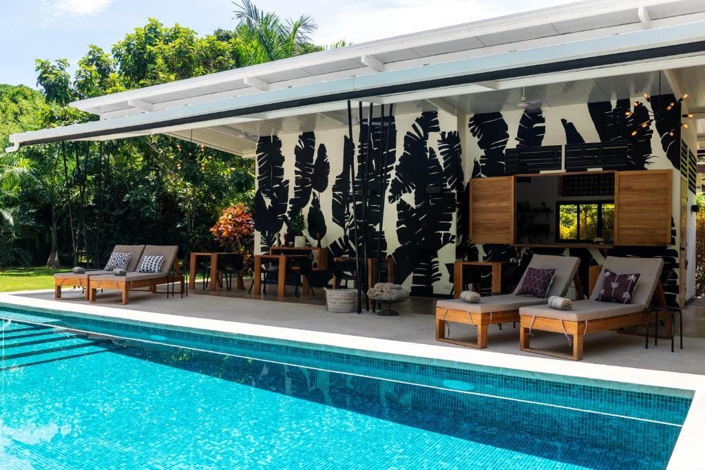 A pool in the foreground with loungers, tables, and a small, tropically-decorated building in the background. 